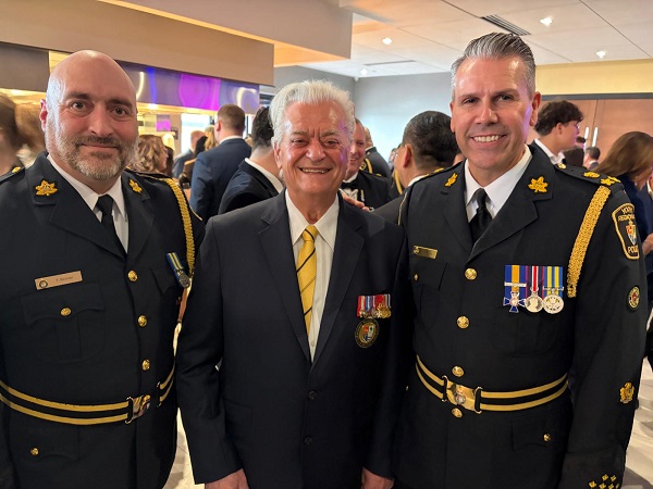 Three men posing together at a formal event; two in dress uniforms with medals flank an older man in a suit and yellow tie, all smiling.
