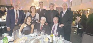 Group portrait of eight adults at a formal banquet, some seated and others standing around a round table with wine bottles and glasses.