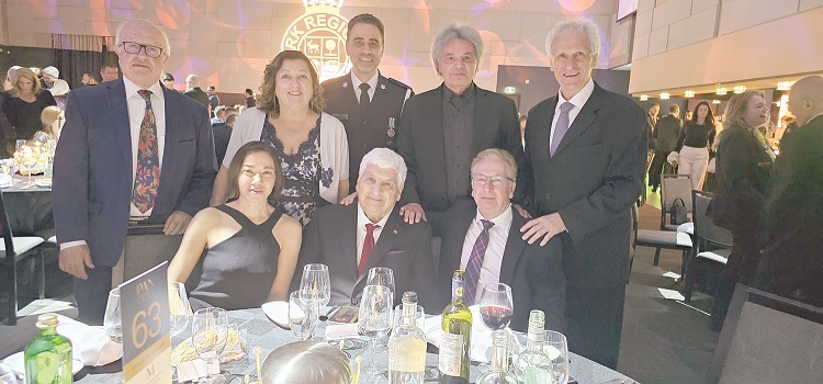 Group portrait of eight adults at a formal banquet, some seated and others standing around a round table with wine bottles and glasses.