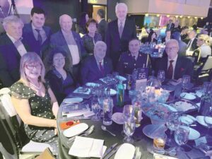 Group of formally dressed guests posed around a banquet table at a gala or reception, with drinks, place settings, and a centerpiece visible.