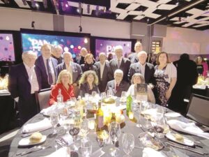 Group of suited and casually dressed adults posing around a round banquet table at a formal gala, with stage screens behind them.