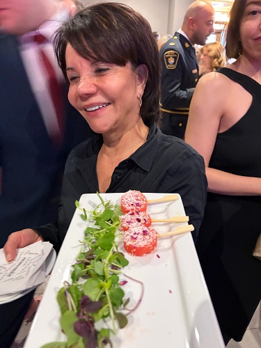 Smiling woman in black holds a long plate of skewered appetizers with greens at a reception.