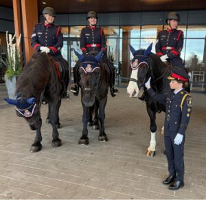 Ceremonial guards on horseback outside a modern building; a young officer stands on the ground beside them.