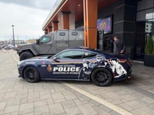 York Regional Police promotional car with 'Investing in Women' graphics parked in front of a modern building; a large armored police vehicle sits behind it.