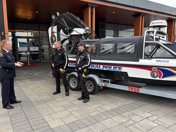 Three officers in black uniforms and a suited supervisor stand by a police boat on a trailer outside a modern glass-front building.