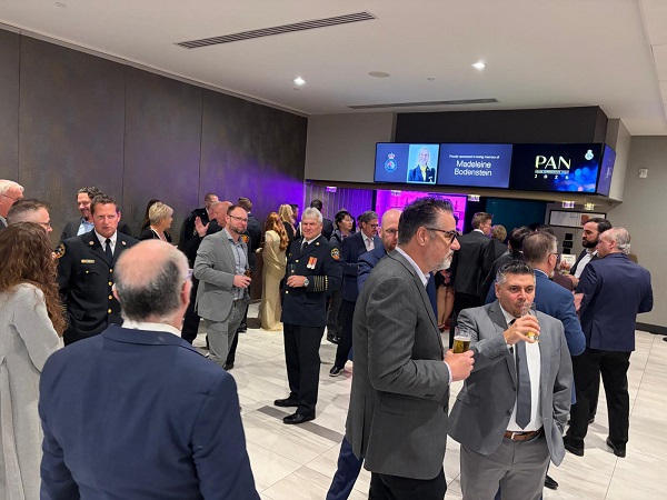 Professionals in formal attire mingling at a reception in a modern lobby, some in uniforms, holding drinks near a cocktail bar with digital signage in the background.