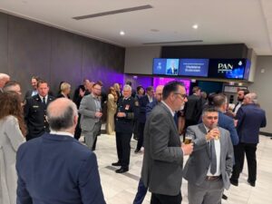 Crowd of professionals in suits and military uniforms mingle at a modern lobby reception, holding drinks with a digital sign in the background.