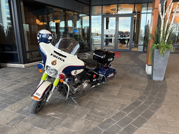 White police motorcycle with blue trim and a helmet on the seat parked outside a glass-front entrance, in a paved area of a modern building.