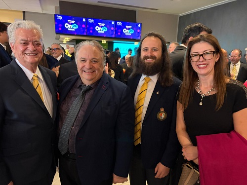 Group of four professionally dressed people posing for a photo at a reception, smiling in a crowded event space with screens in the background, two men in suits with yellow ties and a woman in black with a pink jacket draped over her arm.