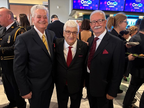 Three men in dark suits posing together and smiling at a formal event, with other guests in the background.
