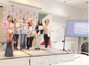 Group of children on a stage holding certificates and gift bags, posing with an adult presenter after a ceremony.