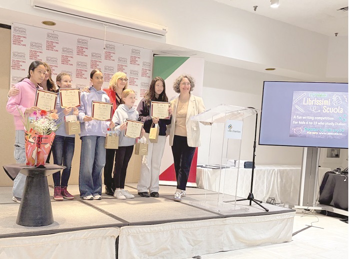 Group of children on a stage holding certificates and gift bags, posing with an adult presenter after a ceremony.