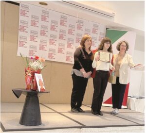 Young girl receives a certificate on stage from two women during an awards ceremony, with sponsor banners in the background.