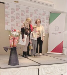 Group of four adults and a boy on a stage posing with a certificate, a flower bouquet on a table to the left.