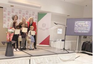 Group of children and a woman posing on a stage after receiving certificates; a bouquet on a table to the left, banners behind, a clear podium, and a screen on the right displaying event info.