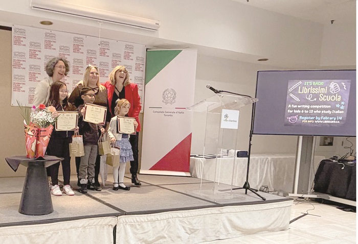 Group of children and a woman posing on a stage after receiving certificates; a bouquet on a table to the left, banners behind, a clear podium, and a screen on the right displaying event info.
