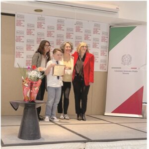 Four people on a stage posing with a certificate award; child center, two women and a woman in red jacket smile beside him, banners and flowers nearby.