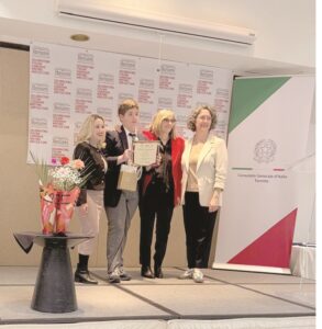 Five people on a stage receiving an award; a person holds a certificate while others smile nearby, with a bouquet on a table and banners in the background.