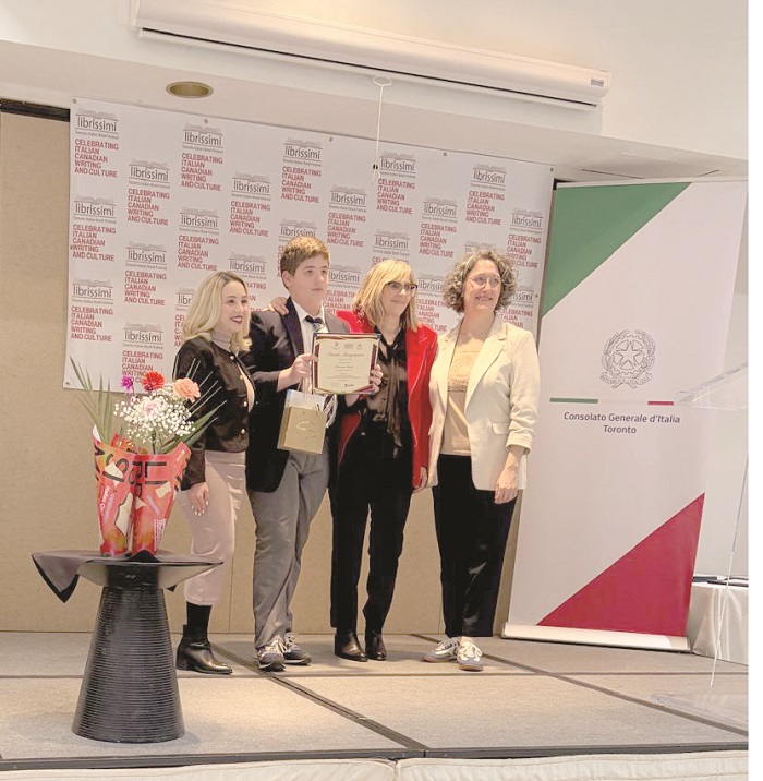 Five people on a stage receiving an award; a person holds a certificate while others smile nearby, with a bouquet on a table and banners in the background.