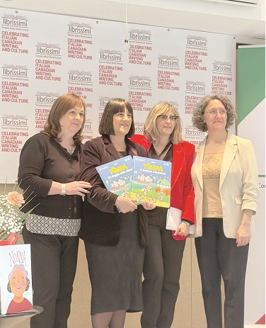 Four women posing at a cultural event; the two center women hold a colorful children's book, with a backdrop of repeated banners about celebrating Italian Canadian culture.