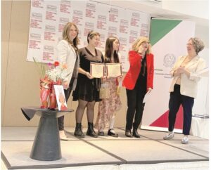 Five women on a stage at an awards ceremony; two hold certificates while a presenter speaks and others smile, with a flower table to the side and a backdrop banner behind them.