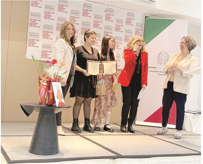 Five women on a stage during an awards ceremony; two hold certificates as a presenter in a red jacket speaks, with banners in the background.