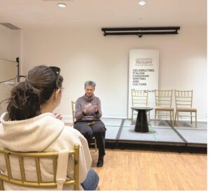 Woman on a small stage speaking to an audience; four chairs behind her and a banner about Italian-Canadian writing and culture.