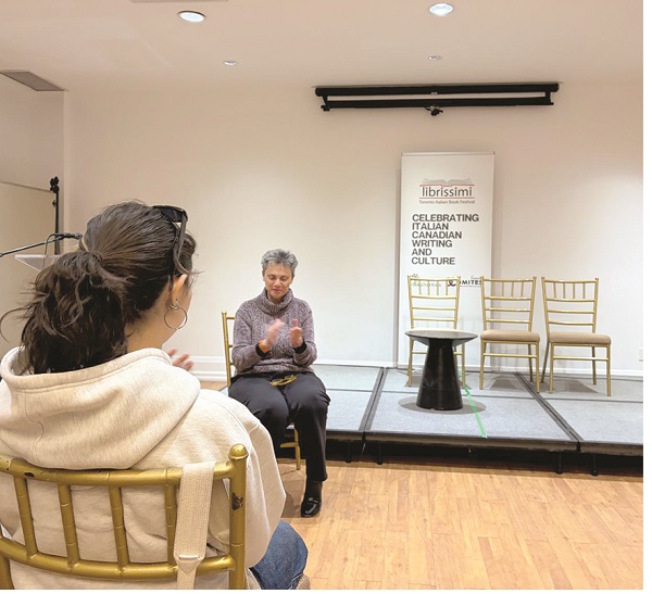 A woman speaks to an audience in a small room, seated on a chair on a raised stage with a banner and four chairs behind her.