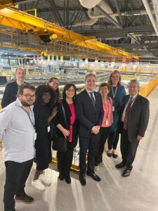 Group of nine professionals posing on a factory floor under a yellow overhead crane.
