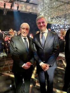 Two men in dark suits pose for a photo at a formal gala, with chandeliers and guests visible in the background.