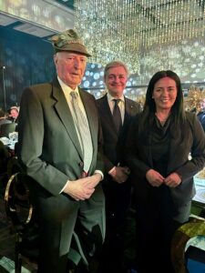 Three people in formal attire pose for a photo at a gala dinner with sparkling hanging lights in the background, a man in a suit wearing a hat on the left.