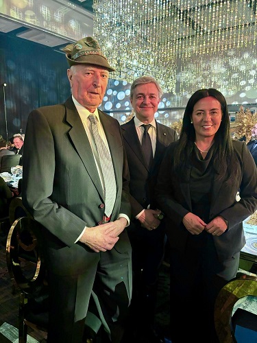 Three people in formal attire pose for a photo at a gala dinner with sparkling hanging lights in the background, a man in a suit wearing a hat on the left.