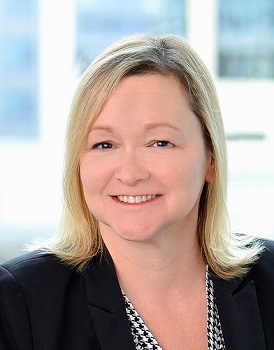 Professional headshot of a smiling blonde woman in a black blazer, facing camera in an office setting, neutral background.