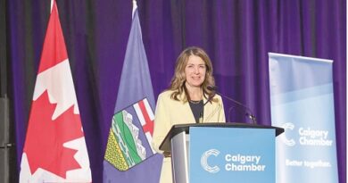 Woman speaks at a podium labeled Calgary Chamber, with Canadian and provincial flags beside her against a purple backdrop.