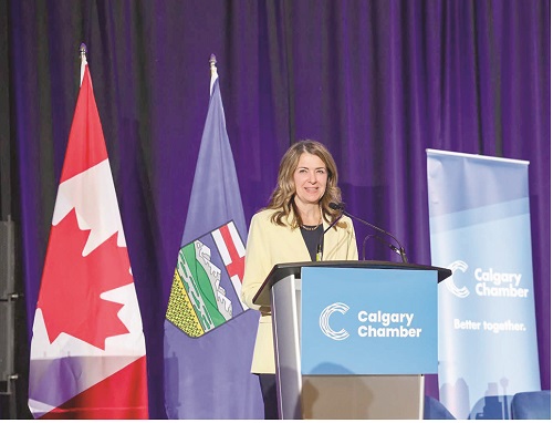 Woman speaks at a podium labeled Calgary Chamber, with Canadian and provincial flags beside her against a purple backdrop.