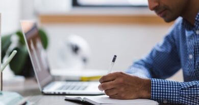 Man in a blue checkered shirt writes in a notebook at a wooden desk with a laptop nearby.