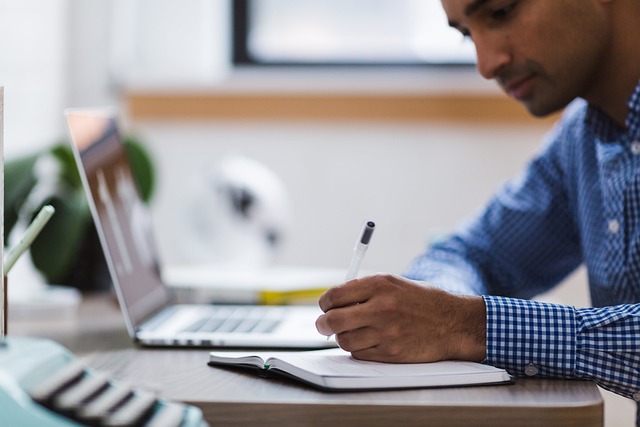 Man in a blue checkered shirt writes in a notebook at a wooden desk with a laptop nearby.
