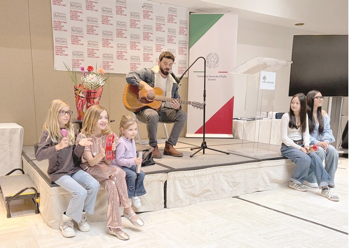 A man sits on stage playing an acoustic guitar while kids sit on both sides, listening. A microphone stand and banners in the background.