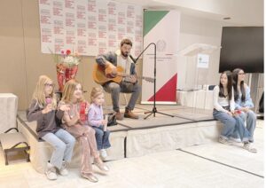 Man with guitar performs on a small stage as several children sit nearby during an indoor event, with banners in the background.