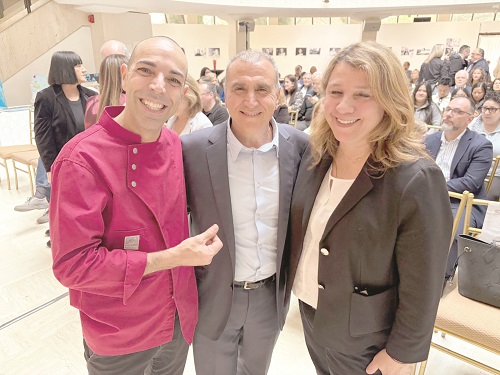 Three people posing for a photo at an event: man in pink shirt left, man in suit center, woman in blazer right, all smiling with a crowd in the background.