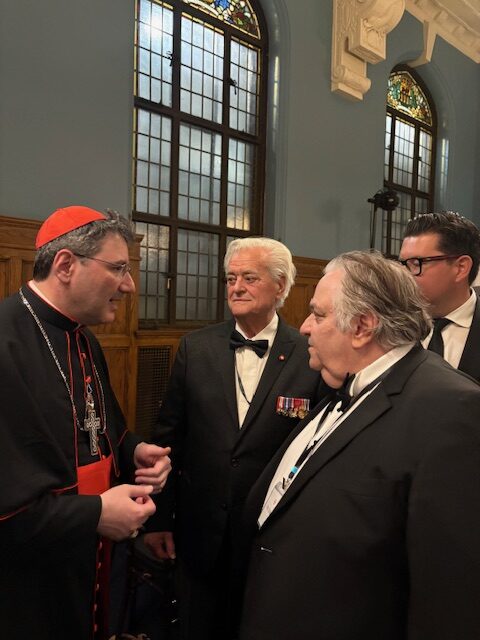 Cardinal in red and black clerical robes chats with two men in black tuxedos in a room with tall stained-glass windows.