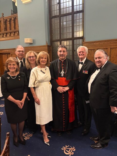 Seven adults in formal attire posing for a group photo in a wood-paneled room; a clergyman in red and black robes stands center.