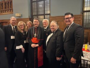 Group of eight adults in formal attire posing for a photo at a reception, with a cardinal in red and black robes center stage in a wood-paneled room.