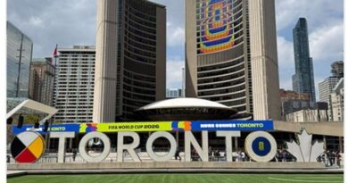 Toronto sign at Nathan Phillips Square with city skyline and banners for FIFA World Cup 2026 behind, showing a green lawn in the foreground and a colorful sculpture spelling TORONTO