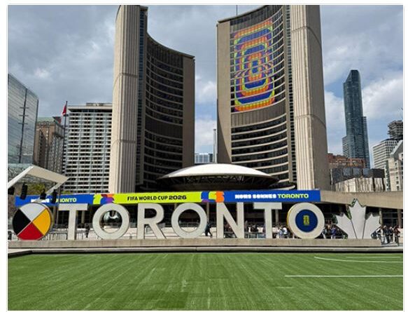 Toronto sign at Nathan Phillips Square with city skyline and banners for FIFA World Cup 2026 behind, showing a green lawn in the foreground and a colorful sculpture spelling TORONTO