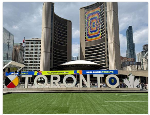 Toronto sign at Nathan Phillips Square with city skyline and banners for FIFA World Cup 2026 behind, showing a green lawn in the foreground and a colorful sculpture spelling TORONTO