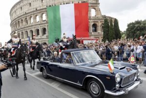 Flag of Italy draped over the Colosseum during a parade with a vintage blue convertible and mounted guards in ceremonial uniforms nearby, while a crowd watches.