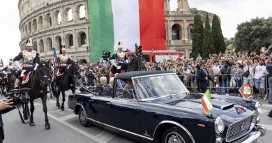 Flag of Italy draped over the Colosseum during a parade with a vintage blue convertible and mounted guards in ceremonial uniforms nearby, while a crowd watches.