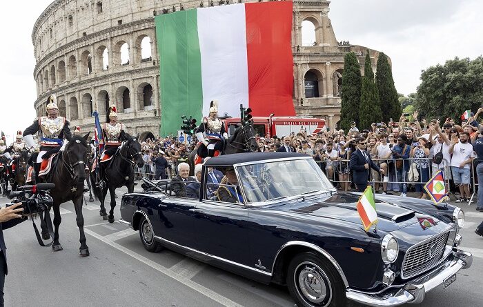 Flag of Italy draped over the Colosseum during a parade with a vintage blue convertible and mounted guards in ceremonial uniforms nearby, while a crowd watches.
