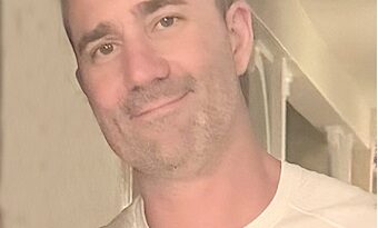 Smiling man with short dark hair and light stubble, wearing a white crew-neck shirt, looking at the camera indoors.
